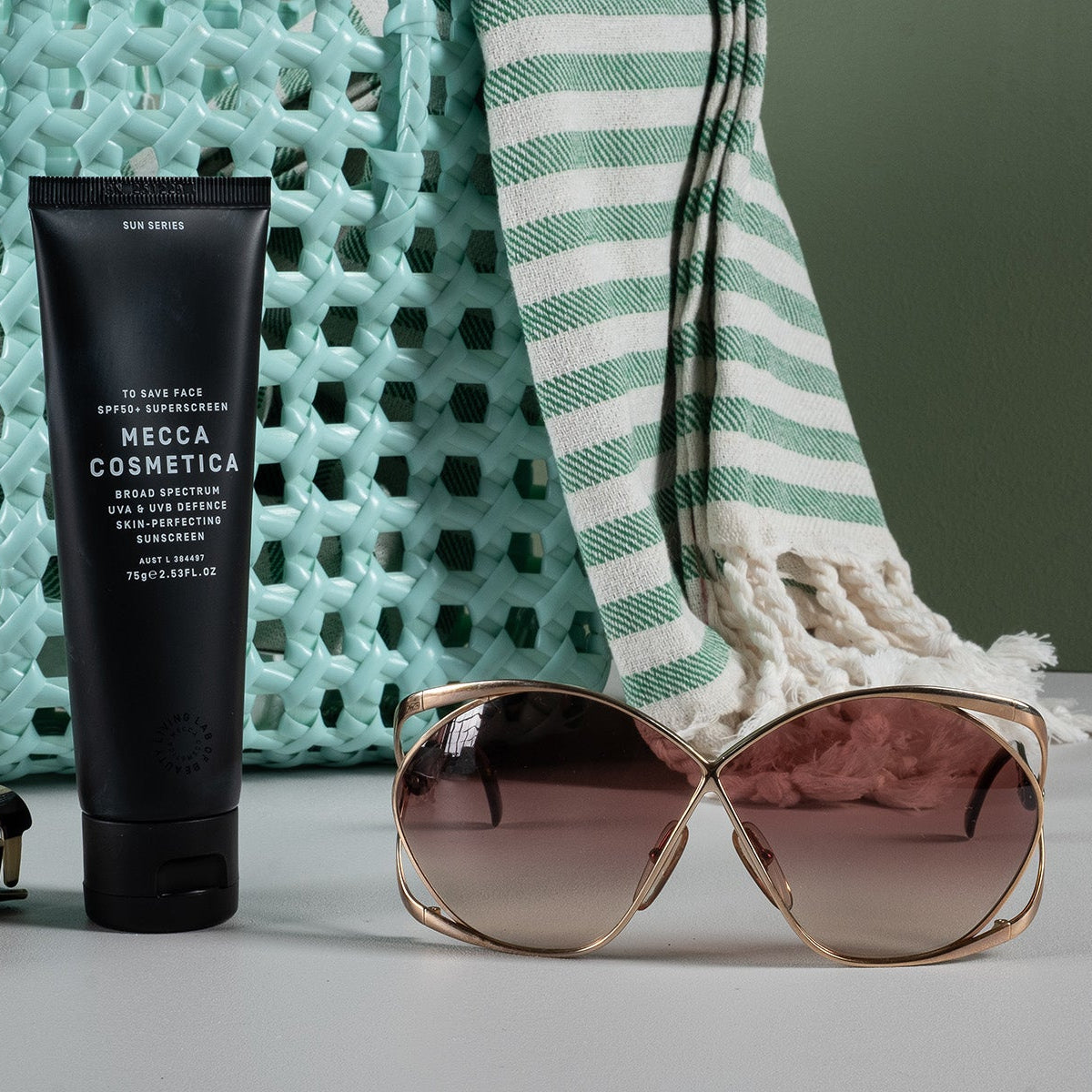 Black tube of Mecca Cosmetica, sunglasses, and a checkered bag on a light surface with a textured green background.